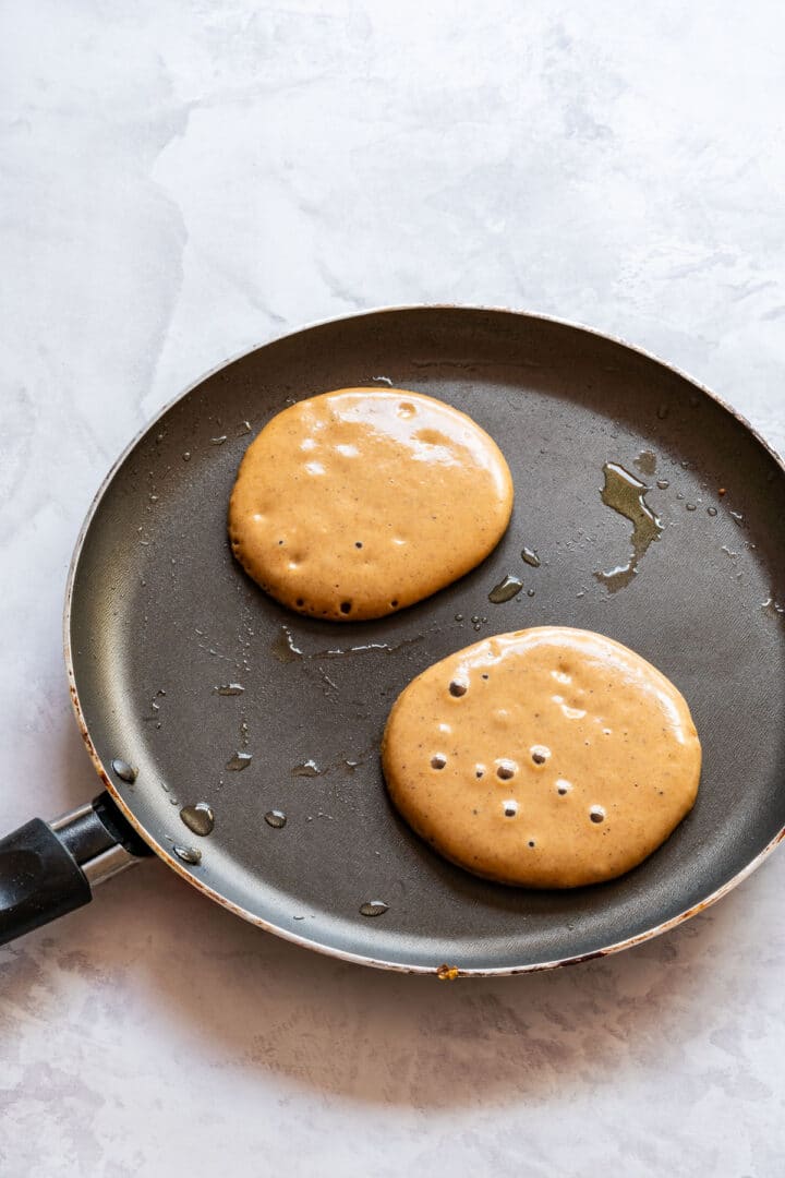 The image shows two pancakes cooking in a frying pan with bubbles forming on the top.
