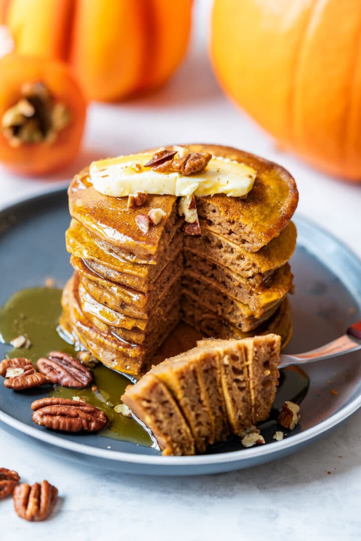 A stack of pumpkin spice pancakes with slice taken out, topped with butter, pecans and honey on a dark plate. Whole pumpkins in the background.