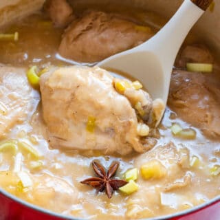 A spoon lifting a chicken thigh from a pot with leeks and star anise in a thick broth.