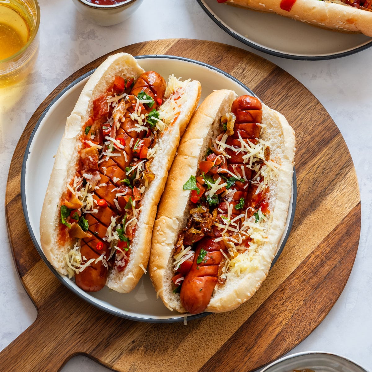 Two gourmet hot dogs in buns topped with shredded cheese, caramelized onions, tomatoes, herbs and sliced chili.