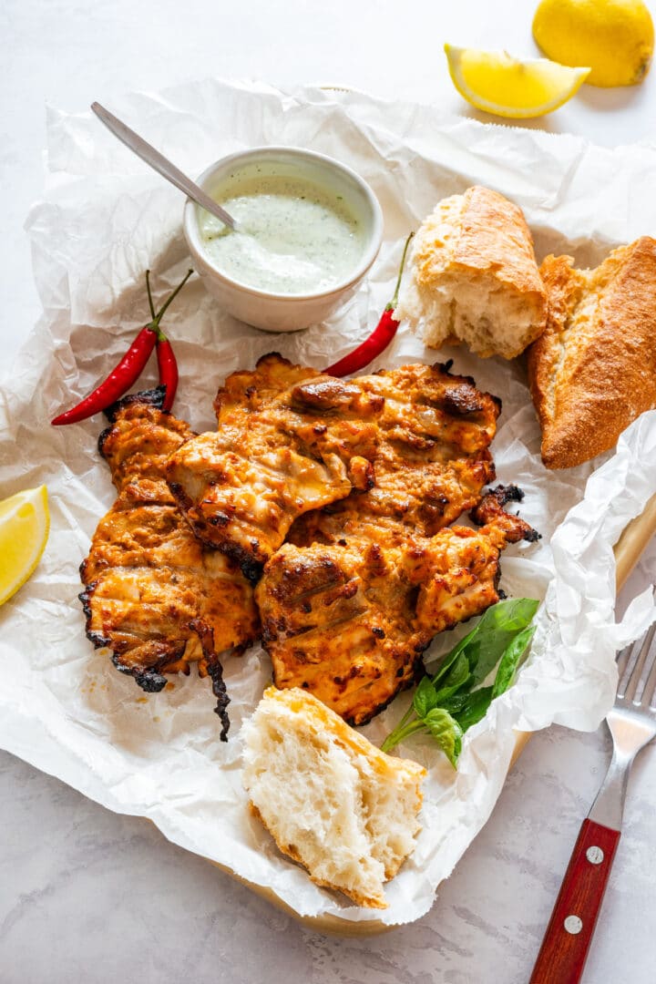 Grilled chicken thighs served with dipping sauce, chili peppers and crusty bread, garnished with lemon and basil.