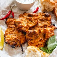 Top-down view of grilled boneless chicken thighs with basil, lemon wedges and chili peppers on a parchment-lined plate.