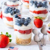Layered no-bake cheesecake cup with blueberries, cream cheese filling and strawberry sauce, styled with fresh fruit and mini American flag for the 4th of July.