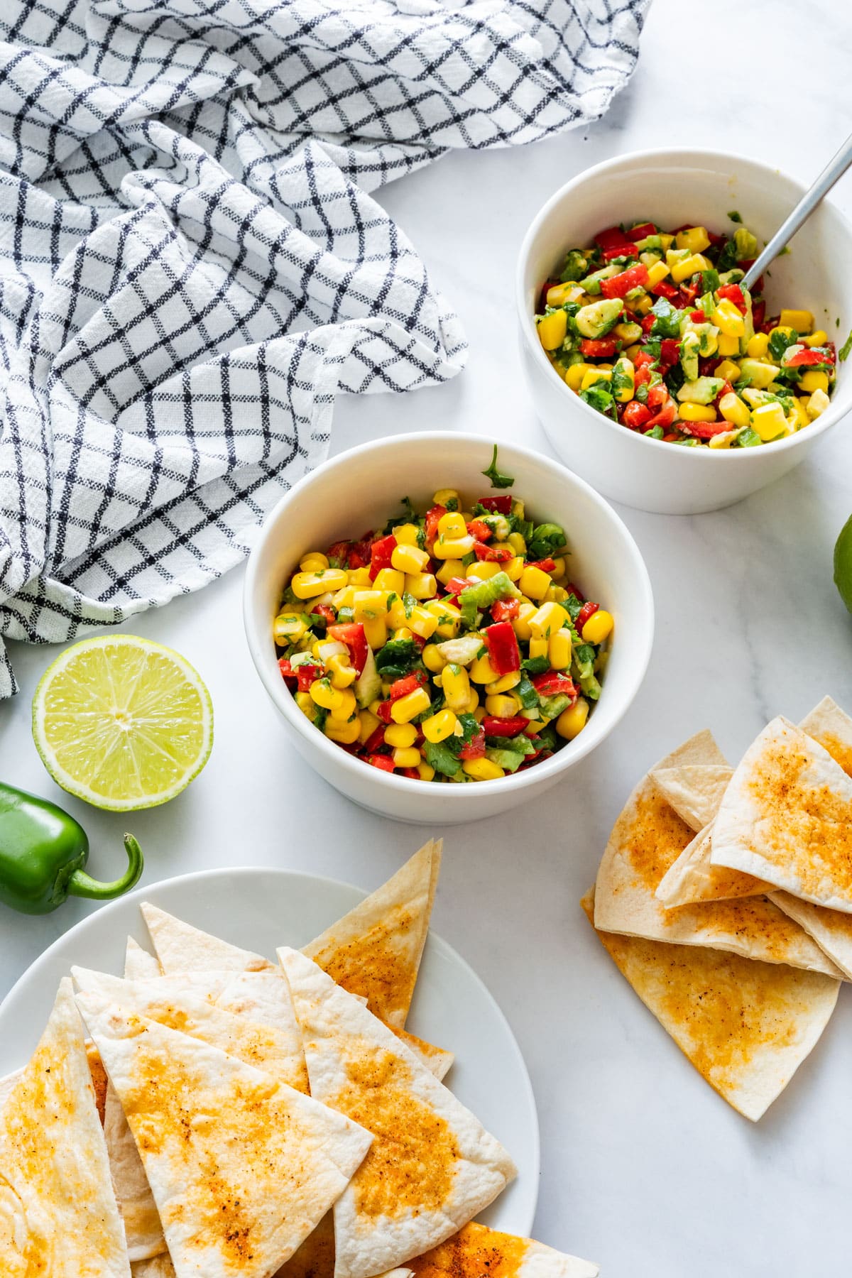 Two white bowls filled with vibrant salsa next to lime, jalapeño and homemade tortilla chips on a marble surface.