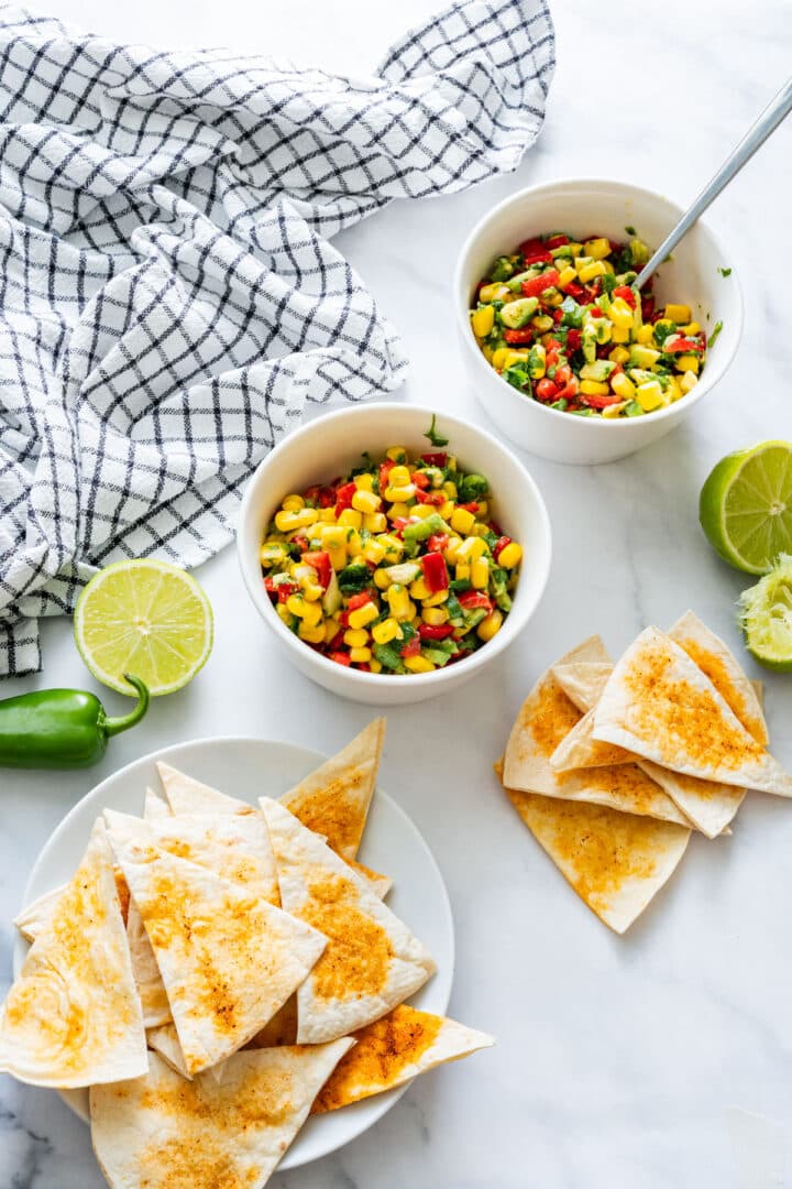 Two bowls of colorful vegetable salsa with lime halves and seasoned tortilla chips on the side.