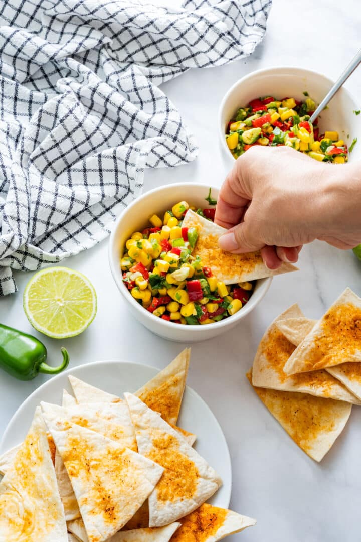 Hand dipping a seasoned tortilla chip into a bowl of vegetable salsa surrounded by lime and peppers.