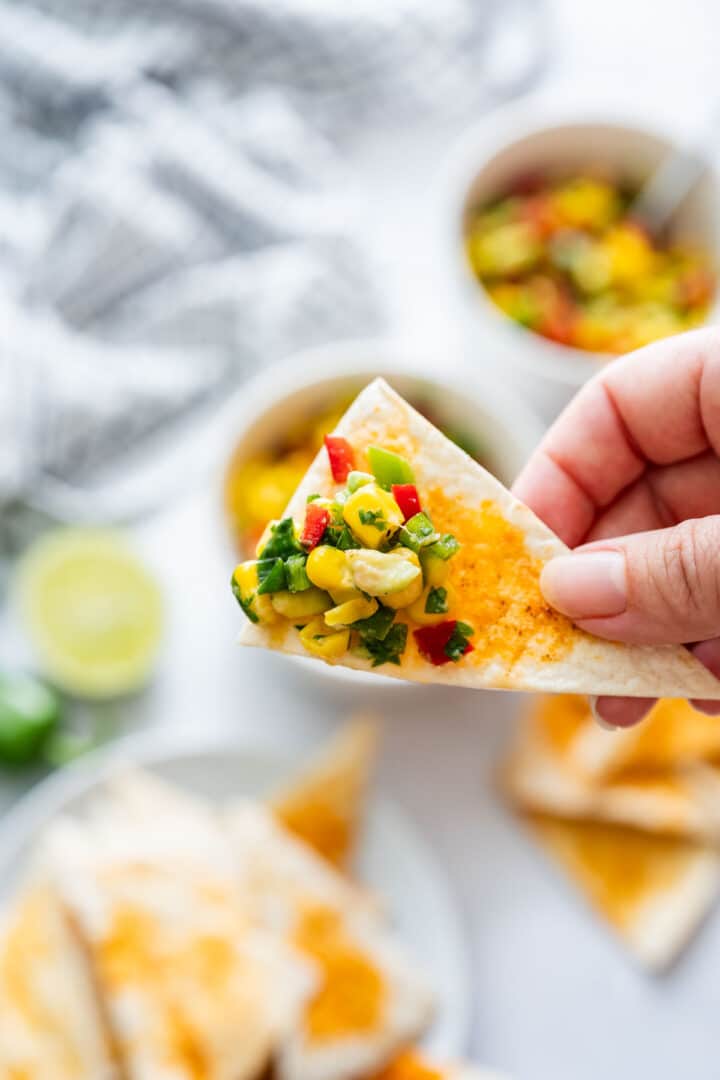Close-up of a chip topped with avocado, corn, peppers and cilantro held above a plate of tortilla wedges.