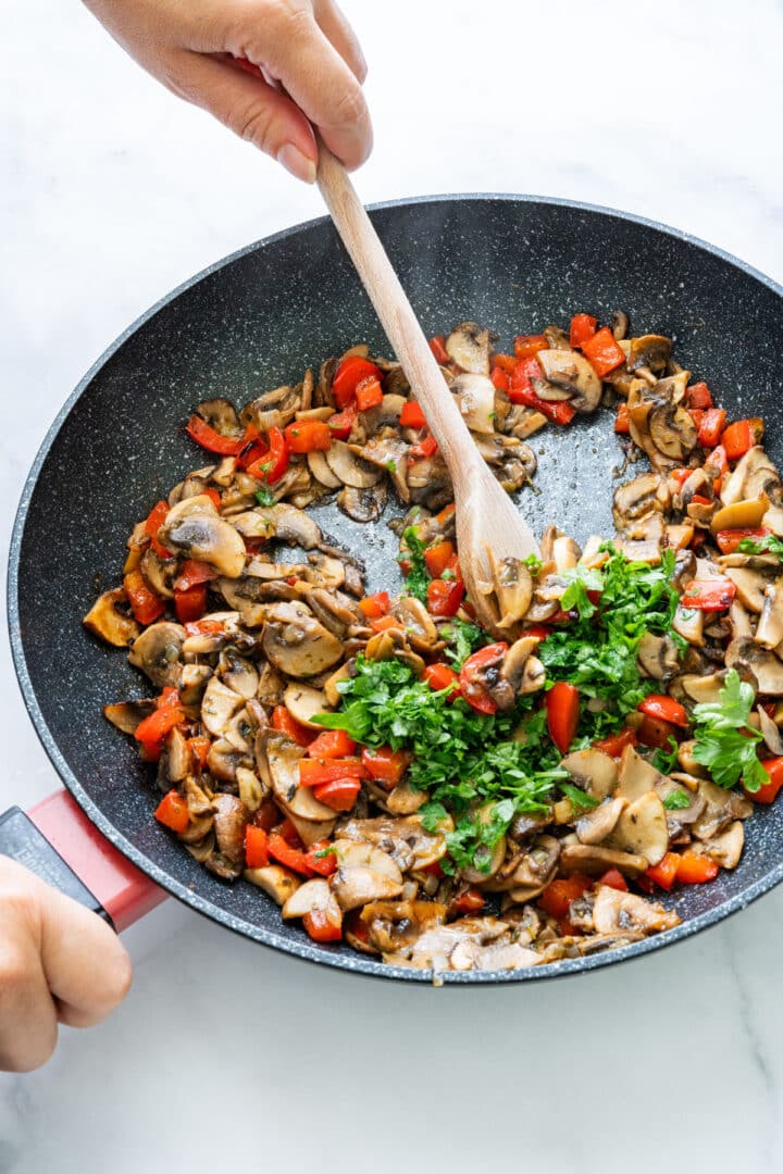 Mushrooms and red peppers being sautéed in a pan with herbs and a wooden spoon.