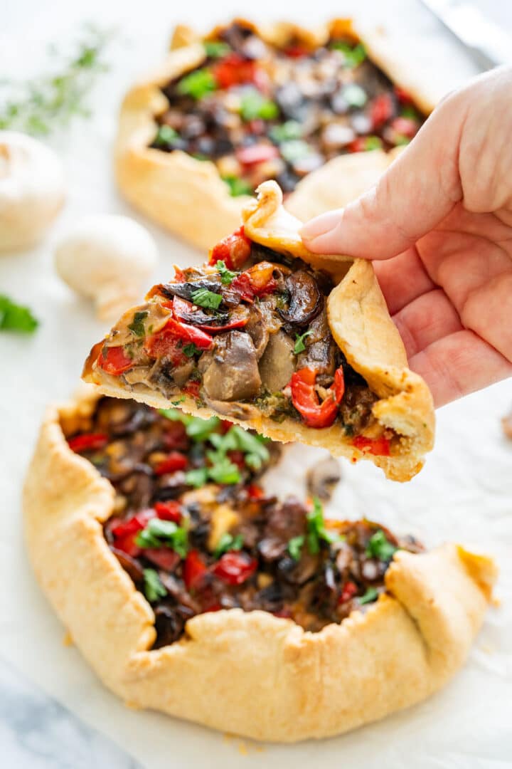 Hand holding a slice of homemade savory galette with mushrooms and peppers, ready to eat.