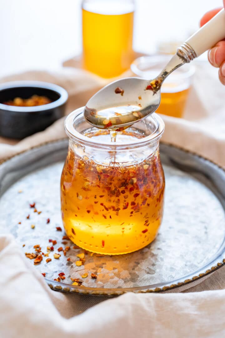 Spoon drizzling hot honey from a jar with visible chili flakes.