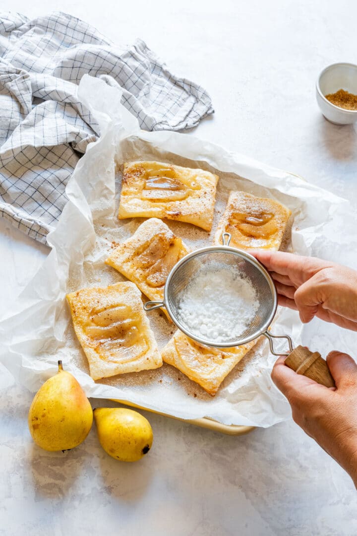 Dusting baked pear tarts with powdered sugar using a small sieve.