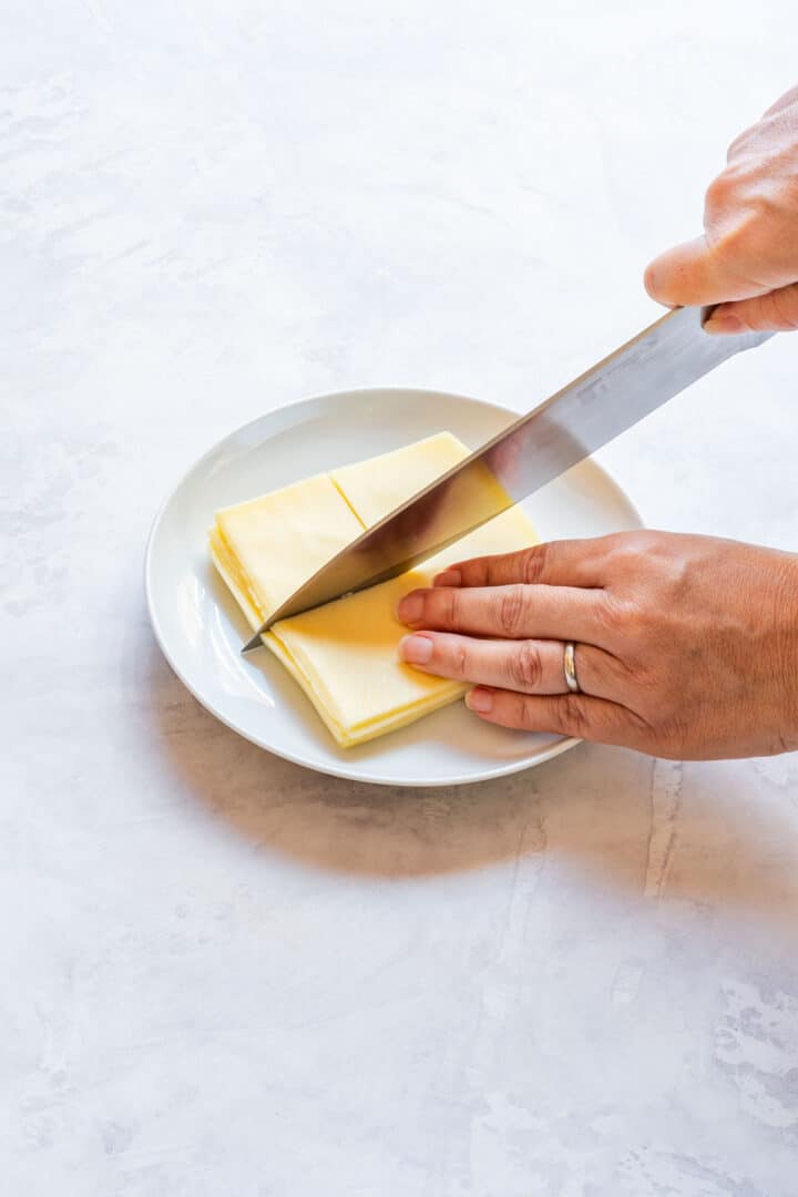 Hand cutting stacked cheese slices into smaller pieces on a white plate.