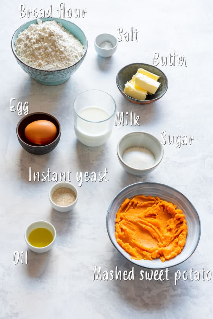 Ingredients for sweet potato dinner rolls, including flour, mashed sweet potato, egg, milk, butter, yeast, sugar, salt and oil with labels.