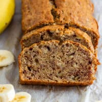 Close-up of a moist slice of simple banana bread with the loaf behind it and banana slices nearby.