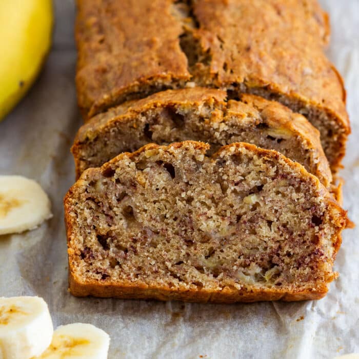 Close-up of a moist slice of simple banana bread with the loaf behind it and banana slices nearby.