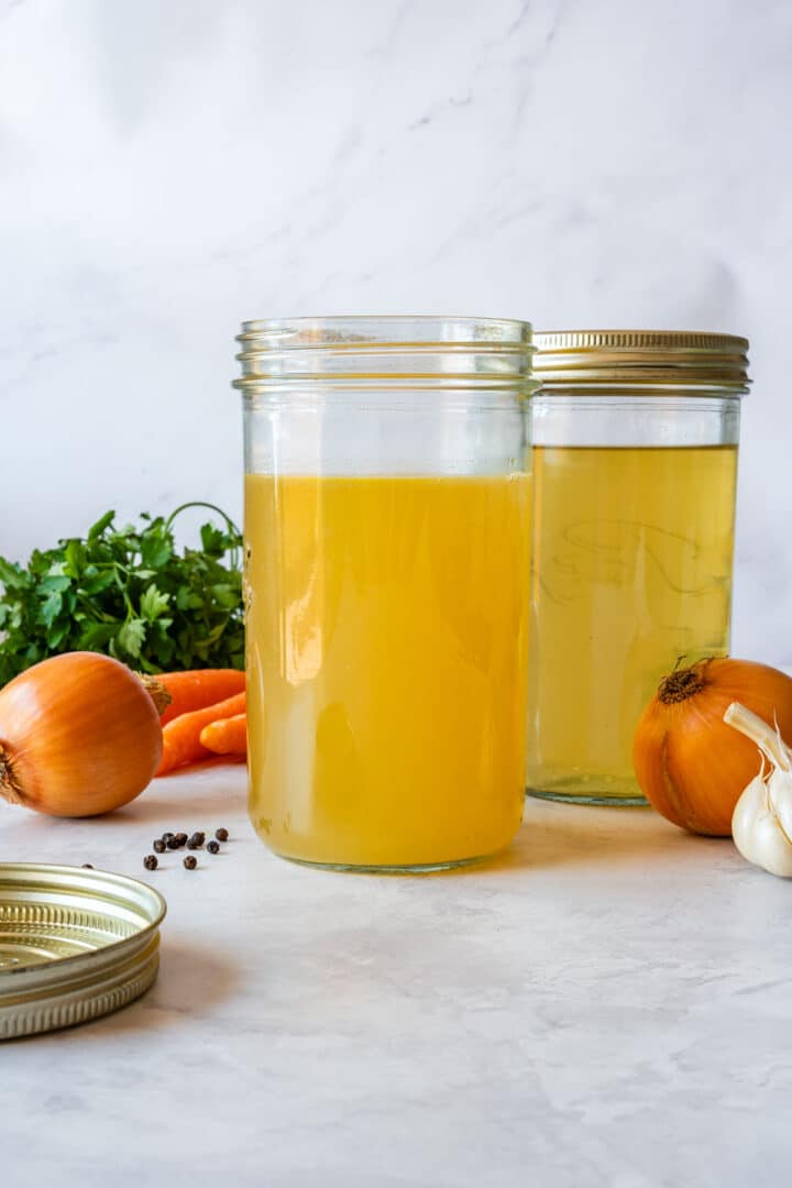 Homemade stock in glass jars with onions, carrots and parsley nearby.