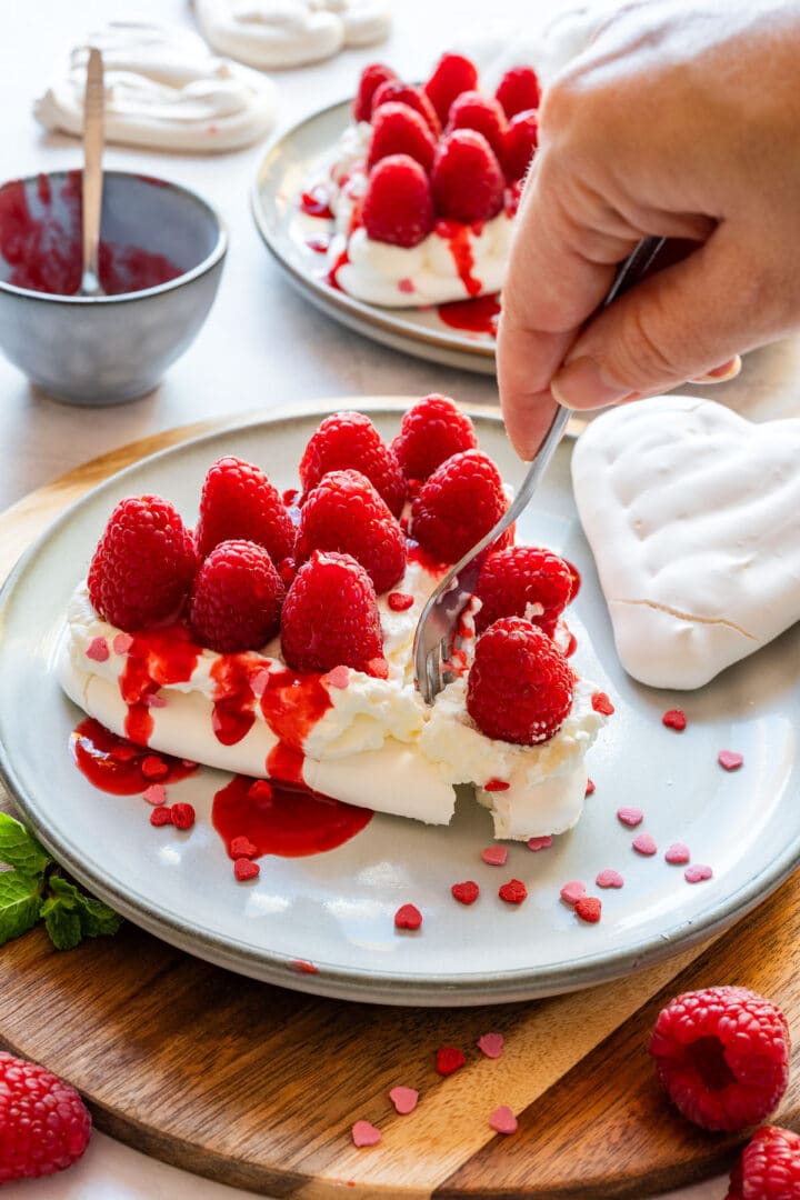 Cutting into a pavlova heart topped with whipped cream, raspberries and raspberry coulis.