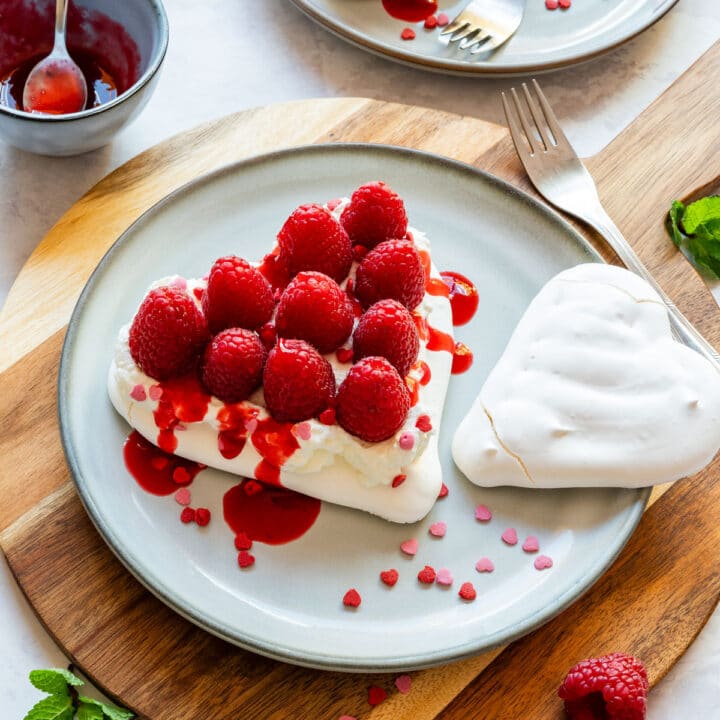 Heart-shaped mini pavlova topped with whipped cream, raspberries, raspberry coulis and sprinkles.