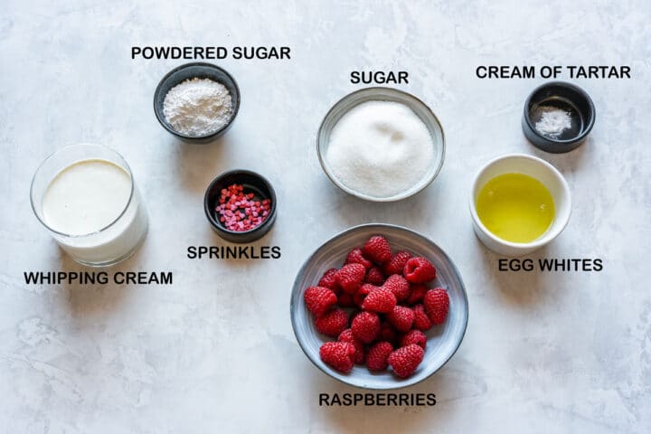 Ingredients laid out for pavlova hearts: egg whites, sugar, cream of tartar, raspberries, whipping cream, powdered sugar, sprinkles.