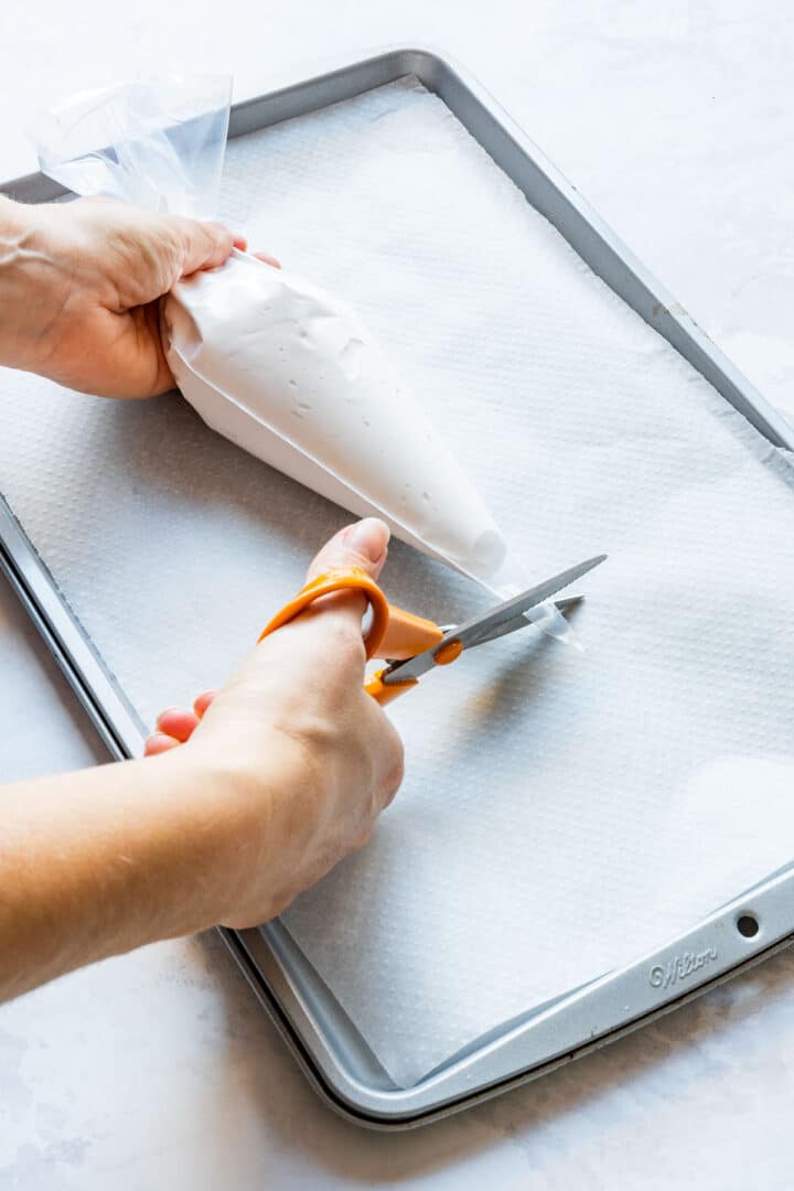Cutting the tip off a piping bag filled with meringue over a parchment-lined baking sheet.