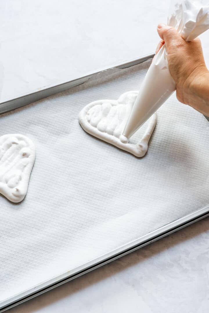 Piping meringue to fill a heart outline on parchment paper.