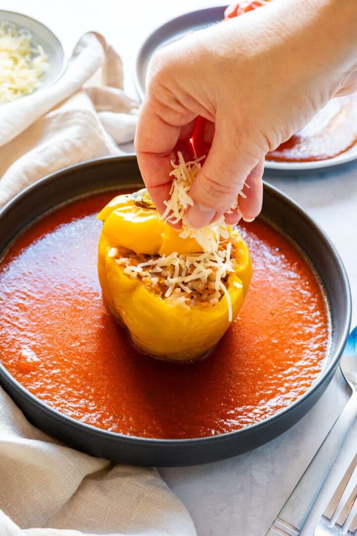 Sprinkling shredded cheese over a stuffed yellow bell pepper in tomato sauce.