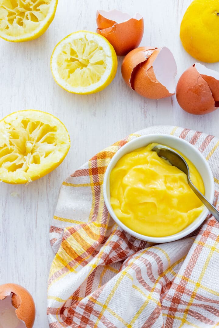 Bowl of lemon curd with spoon, lemon halves and eggshells on a light surface.