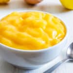 Creamy whole egg lemon curd in a white bowl with a spoon on a wooden table.