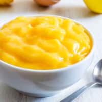 Creamy whole egg lemon curd in a white bowl with a spoon on a wooden table.