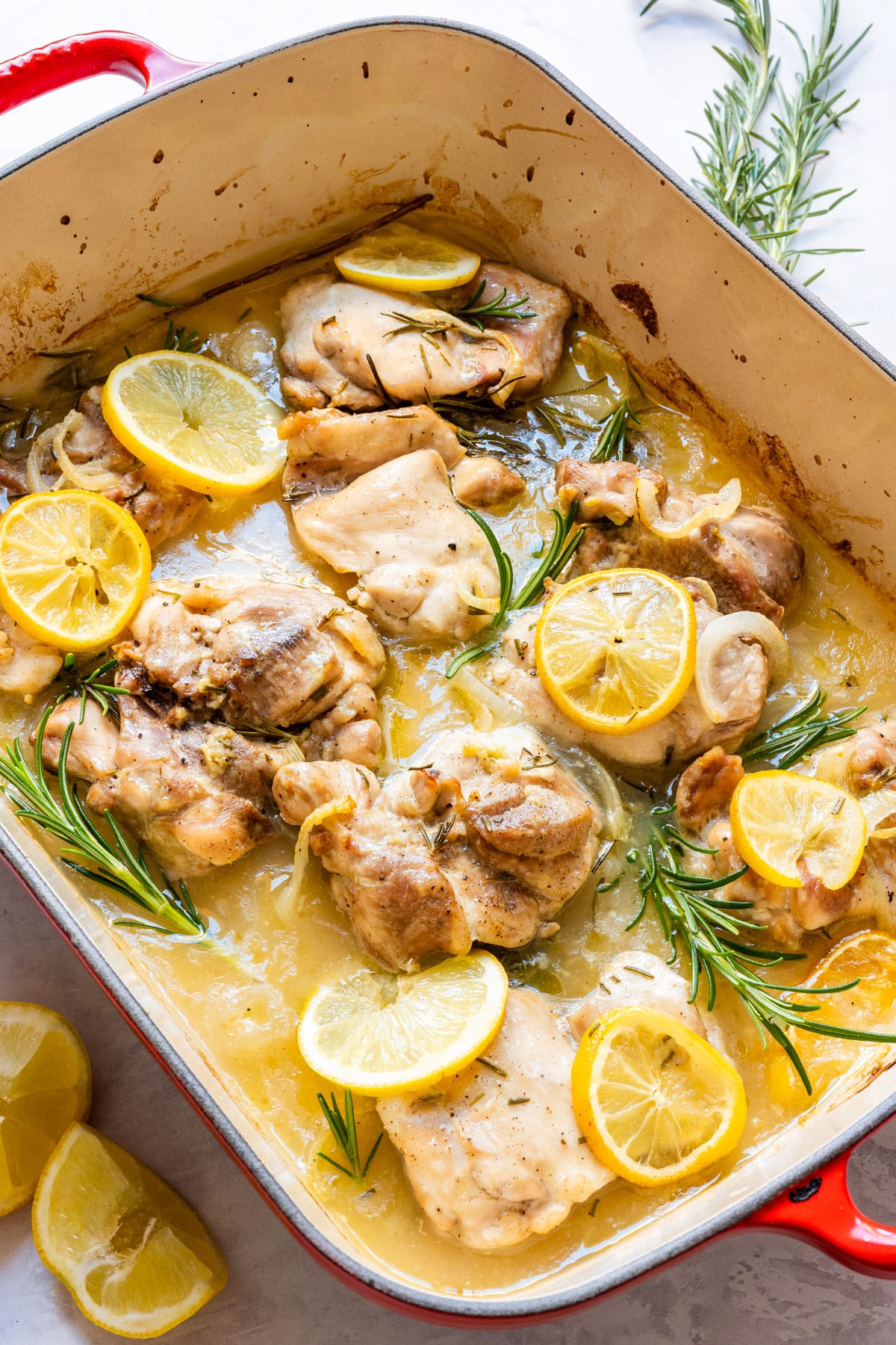 Lemon rosemary chicken thighs baked with lemon slices and rosemary in a red baking dish.