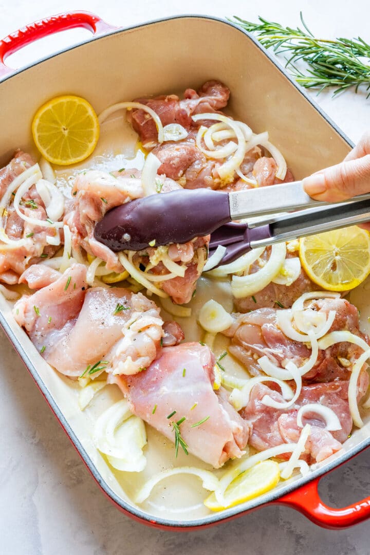Tossing chicken thighs with sliced onion, garlic and lemon in a baking dish using kitchen tongs.