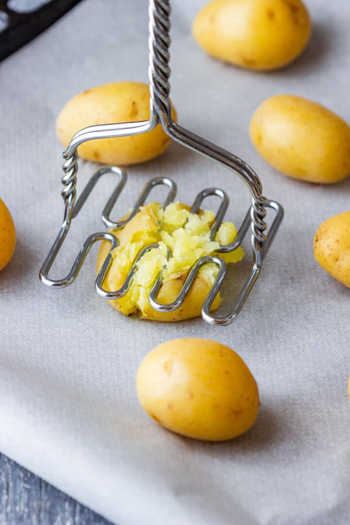 Baby potato being flattened with a potato masher on a parchment-lined baking sheet.