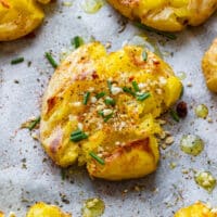 Golden garlic smashed potatoes on parchment paper with herbs, chives and olive oil.