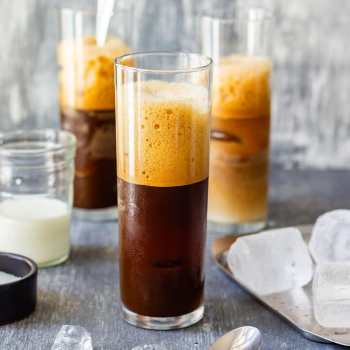 Glass of Greek frappe with a thick foam top, with a small jar of milk and ice beside it.