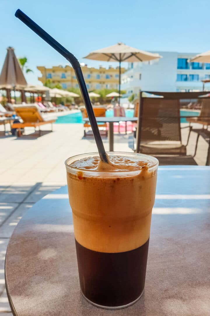 Greek frappe in a tall glass with a straw on a poolside table, with sun loungers and umbrellas in the background.