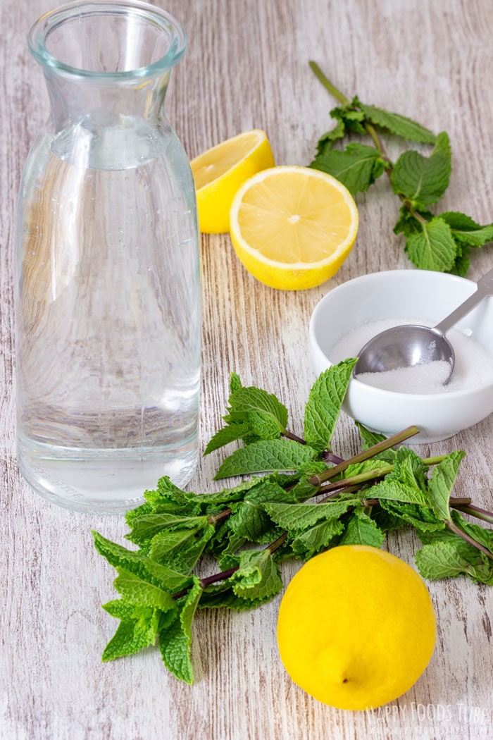 Ingredients for mint lemonade: a carafe of water, fresh lemons, mint sprigs and a small bowl of sugar.