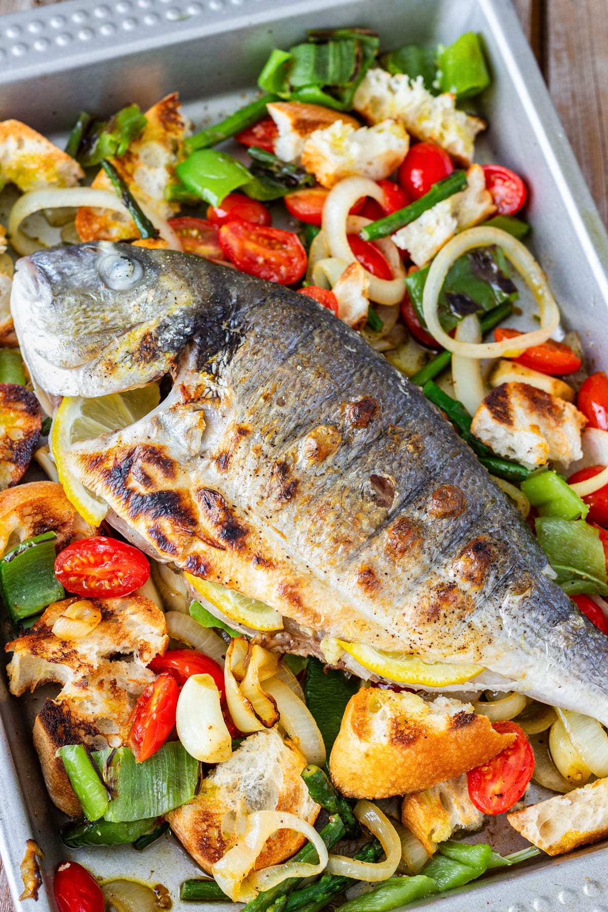 Grilled whole sea bream with crispy skin on a tray of bread salad with cherry tomatoes, onion, green pepper and grilled bread.