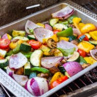 Colorful grilled vegetables in a metal tray on a charcoal grill.
