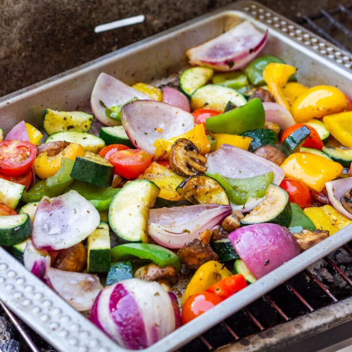 Colorful grilled vegetables in a metal tray on a charcoal grill.