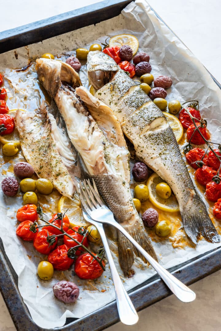 Roasted whole fish opened on a baking sheet with tomatoes, olives, lemon slices and pan juices.