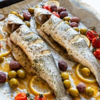 Close-up of two whole roasted branzino with mixed olives, roasted cherry tomatoes, lemon slices and golden pan juices.