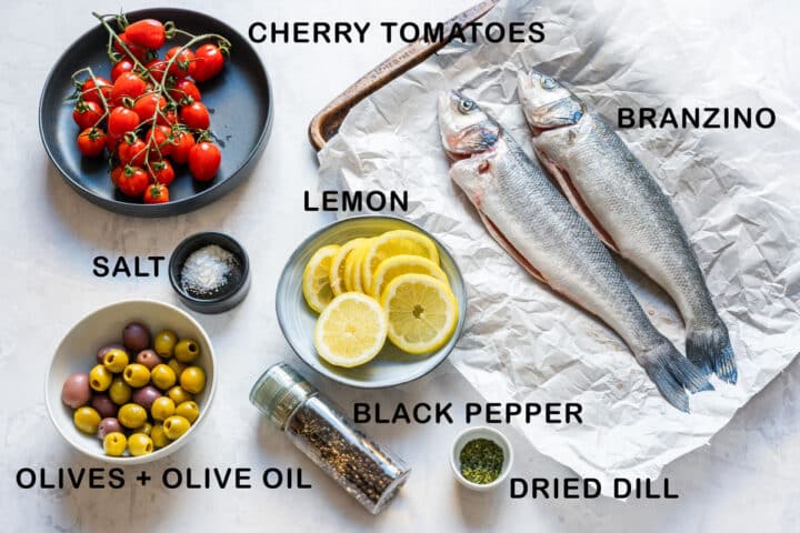 Ingredients for whole roasted branzino, including two raw fish, cherry tomatoes, lemon, mixed olives, salt, black pepper and dried dill.
