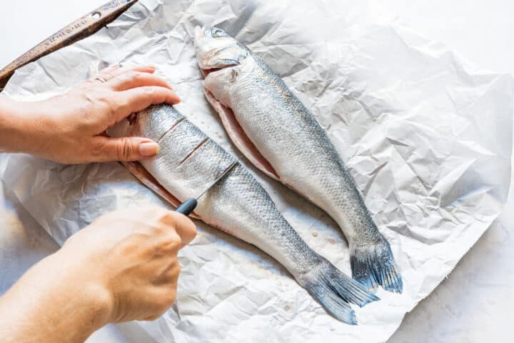 Making shallow diagonal cuts in a raw branzino with a second fish alongside on parchment paper.