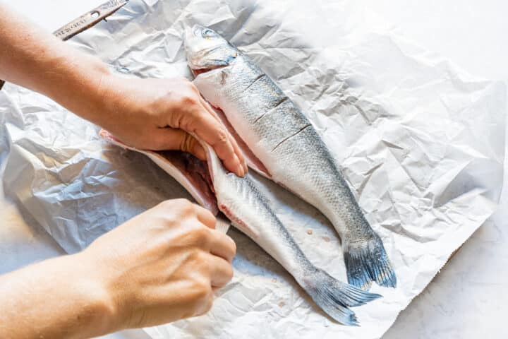Opening the belly of a cleaned whole fish before seasoning, with another scored fish beside it.