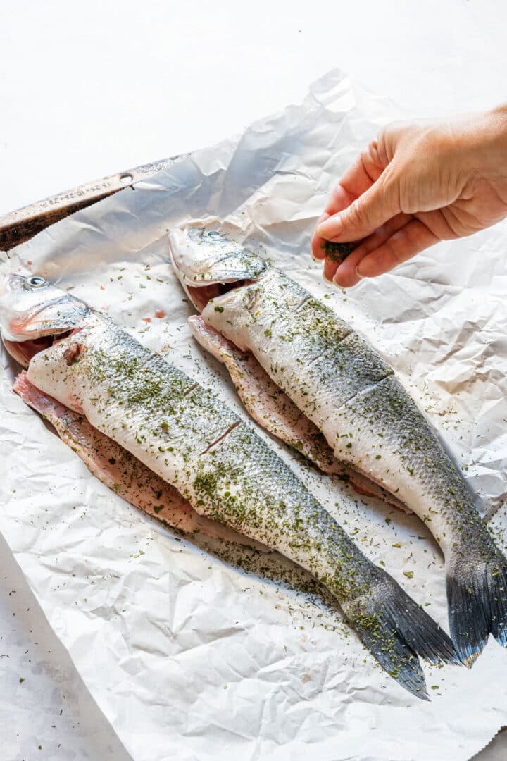 Sprinkling dried dill over two scored whole branzino on parchment paper.
