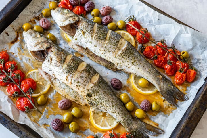 Overhead view of roasted branzino with mixed olives, blistered cherry tomatoes, lemon slices and pan juices.
