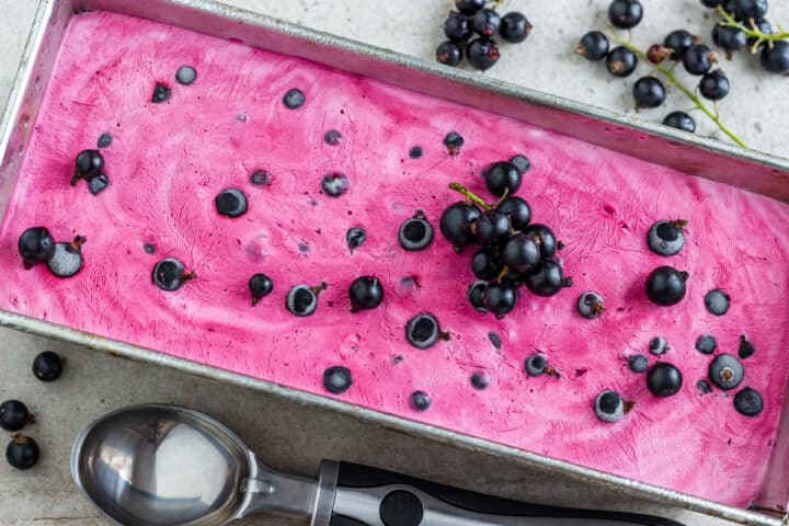 Overhead view of smooth pink berry ice cream in a loaf pan, topped with frozen blackcurrants and a small cluster of berries.