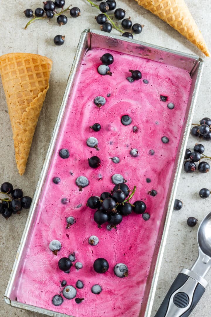 Loaf pan of just-frozen pink currant ice cream scattered with dark berries and framed by two waffle cones.