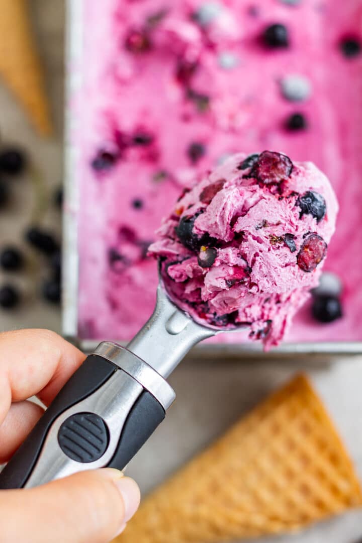 A hand holding a scoop of pink blackcurrant ice cream with whole berries and a waffle cone below.