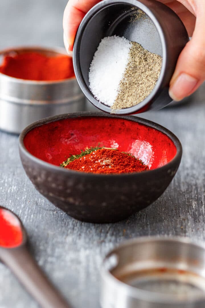 Salt and black pepper being tipped into a bowl with paprika and dried dill.